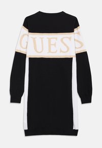 Čierno-biely pletený sveter s dlhým rukávom a zlatým logom "GUESS" na hrudi. Rukávy sú dlhé, manžety sú rebrované a po stranách sú rozparky.