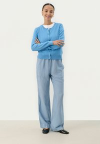 Cardiganazzurro chiaro con scollatura rotonda e bottoni, abbinato a pantaloni larghi azzurri chiari. Ballerine nere e una t-shirt bianca sotto.