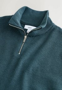 Maglione a costine di un verde petrolio scuro con colletto con zip. Presenta una zip argentata e una texture intrecciata. L'etichetta indica la taglia Media.