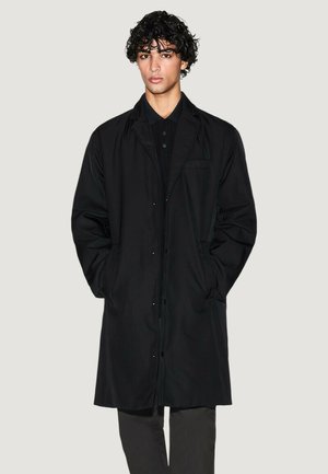 Cappotto lungo nero con chiusura a bottoni, tasche laterali e una vestibilità leggermente oversize. Realizzato in un tessuto liscio e resistente.
