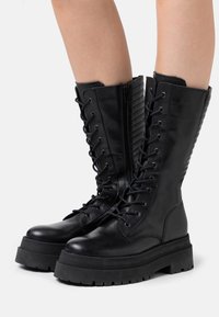 Bottes en cuir noir à lacets avec une semelle en caoutchouc épaisse et des détails matelassés sur les côtés, équipées d'une fermeture éclair latérale pour un enfilage facile.