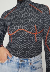 Svart långärmad topp med vit zigzag-mönster och orangea sömmar, med en hög halsdesign och åtsittande silhuett. Kombinerad med denimjeans.