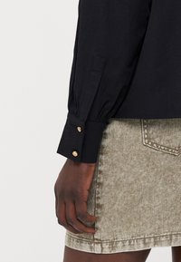 Black long-sleeve shirt with puffed sleeves, featuring gold button details at the cuffs, paired with a beige textured skirt.