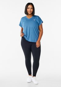 Blauw atletisch shirt met korte mouwen en een v-hals, gecombineerd met zwarte leggings en witte sneakers op een neutrale achtergrond.