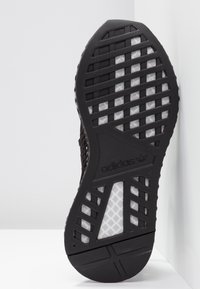 Czarna gumowa podeszwa buta z teksturowanym wzorem i białymi akcentami. Zawiera logo, rowki w kształcie siatki oraz podniesioną krawędź dla lepszej przyczepności.