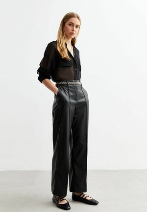 Camisa negra semitransparente de botones con mangas largas, combinada con pantalones de cuero sintético negros de talle alto y pierna ancha, y zapatos planos con correas.