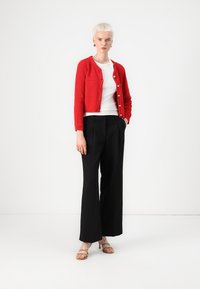Un cardigan en tricot rouge sur une chemise blanche et un pantalon large noir. La tenue comprend des sandales beige clair et présente des boutons dorés sur le cardigan.