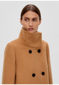 Manteau en laine de chameau avec un col haut et un design à double boutonnage. Comprend de grands boutons noirs et une finition douce et texturée.