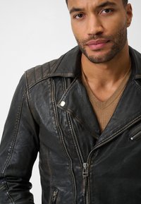 Schwarze Leder-Motorradjacke mit gesteppten Schultern, silbernen Beschlägen und asymmetrischen Reißverschlussdetails, getragen über einem braunen V-Ausschnitt-Pullover.