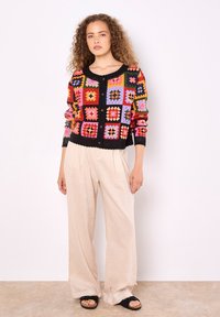 Bunte gestrickte Strickjacke mit einem Patchwork-Design aus Quadraten in Rot, Pink, Blau und Gelb; schwarze Besätze und Knöpfe. Getragen mit beigen, weiten Hosen.