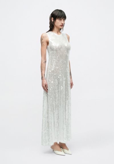 Hosszú, ujjatlan, áttetsző világosszürke ruha flitterekkel, egyenes szabással és finom textúrával. Fehér, hegyes orrú cipővel viselve.