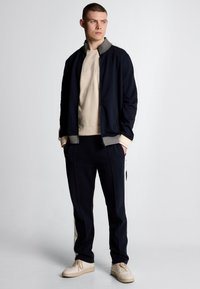 Tuta della marina con accenti grigi, composto da giacca con zip, pantaloni abbinati e sneakers beige. Tessuto liscio con un design aderente.