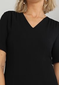 Svart klänning med korta ärmar och V-ringning. Mjukt, lätt tyg med subtil textur. Inga synliga mönster eller utsmyckningar.