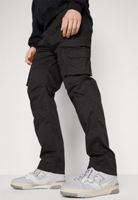 Person som bär svarta cargo-byxor och vita New Balance-sneakers, står med händerna avslappnat vid sidorna mot en vanlig bakgrund.