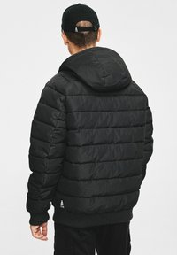 Czarna puchowa kurtka z poziomymi szwami, kapturem i ściągaczami na rękawach. Posiada logo na dole i gładką, matową fakturę.