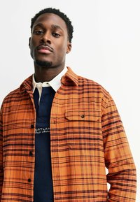 Flanellen overhemd in oranje en zwart geruit patroon, met knoopsluiting, een borstzak en een wit onderhemd met kraag.