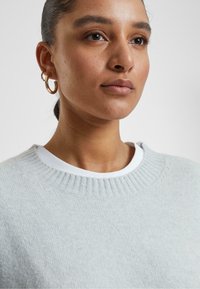 Vaaleanharmaa neulepusero, jossa on ribbireunus, kerrostettuna valkoisen pyöreäkauluksisen paidan päälle. Mallilla on päällään kultaiset rengaskorvakorut, ja hänen ihonsa on sileä ja hiukset tummat.