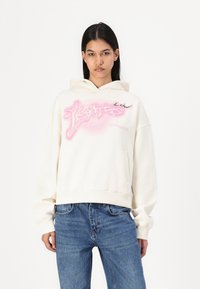 Felpa crema con un design grafico rosa, dotata di tasca frontale, cappuccio con cordino e polsini a coste. Abbinata a jeans blu.