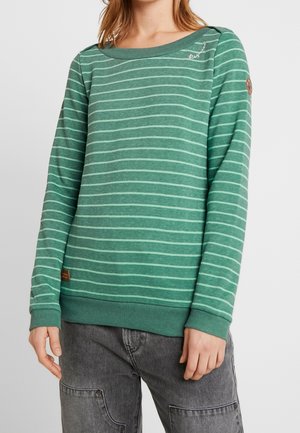 Vrouw die een groene lange mouwen sweatshirt draagt met lichte horizontale strepen en donkergrijze broek, staand tegen een egale achtergrond.