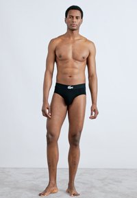 Zwarte slip in briefstijl met een effen ontwerp en een contrasterende witte logo-elastiek. Model poseert rechtop tegen een effen achtergrond.