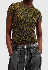 Person som bär en kortärmad topp med grön och svart leopardmönster och löst sittande, blekta svarta jeans med händerna i fickorna.