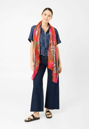 Vrouw draagt een blauw vest met rits, wijde donkere jeans, zwarte sandalen en een lange kleurrijke sjaal met rode, roze en dierenprintpatronen.