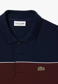 Navy ja burgundi polo särk tekstuuriga viimistlusega, millel on kolme nupu rivi, valge aktsentlint ja roheline tikandiga logo.