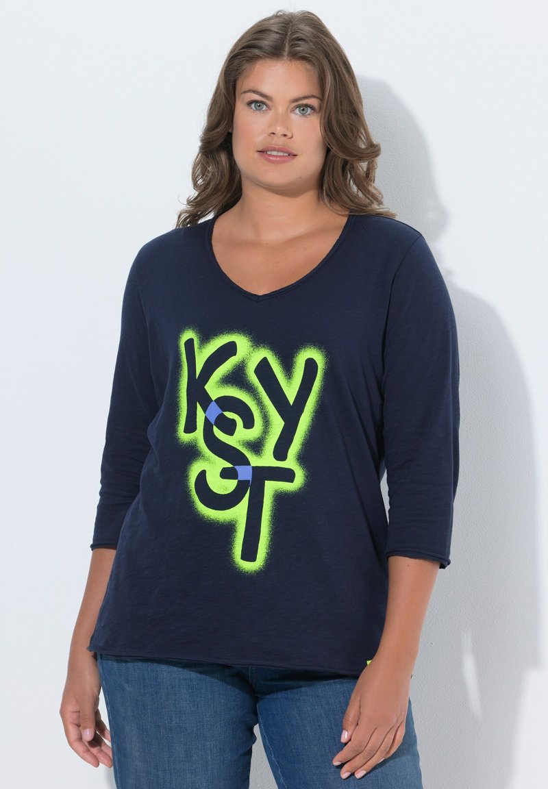 Femme aux cheveux ondulés portant une chemise à manches longues bleu marine avec un texte stylisé vert vif et noir "K S Y S T" et un jean bleu.