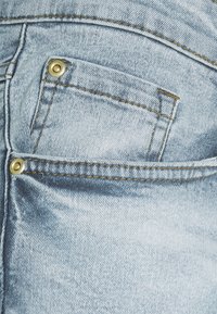 Jeans de denim de color azul claro con un destacado bolsillo trasero con herrajes dorados y costuras doradas a lo largo de las costuras. Textura suave y acabado desgastado.