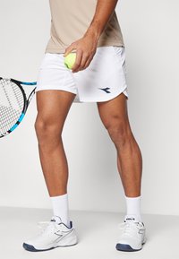 Weiße Sportshorts mit einem blauen Logo, kombiniert mit weißen Sneakers und weißen Sportsocken. Die Person hält einen gelben Tennisball und einen Schläger.