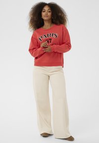 Ženska s kodrastimi lasmi, oblečena v rdečo majico z napisom "Pariz", široke kremne hlače in rjave čevlje, stoji z rokami križanimi.