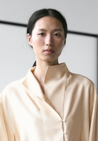 Vrouw met donker haar in een staart draagt een beige shirt met hoge kraag en twee knopen, kijkt recht in de camera tegen een neutrale achtergrond.