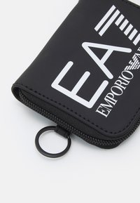 Juodas užtrauktuku uždaromas piniginė, kuriame yra baltas "EA7 Emporio Armani" tekstas ir logo. Lygaus paviršiaus, su prisegtu apvaliu žiedu. Kompaktiška stačiakampio formos.