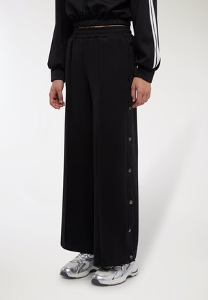 Pantaloni della tuta neri a gamba larga con coulisse in vita e dettagli laterali con bottoni a pressione. Abbinati a scarpe sportive argento. Tessuto dalla texture morbida.