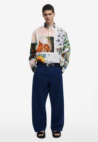 Camicia con bottoni e stampe grafiche miste in colori pastello, abbinata a pantaloni larghi blu notte e sandali neri. La camicia presenta un colletto a punta.