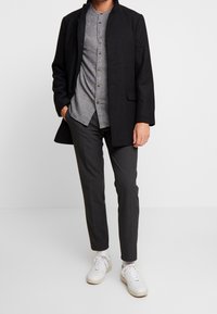 Hombre con abrigo negro, camisa gris abotonada, pantalones gris oscuro y zapatillas blancas, de pie con una mano en el bolsillo.