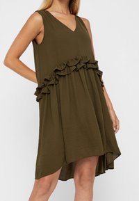 Tummanvihreä hihaton mekko, jossa on V-pääntie. Mekossa on röyhelökoristeet vyötärön ympärillä ja takana hieman pidempi helma. Materiaali on sileä.