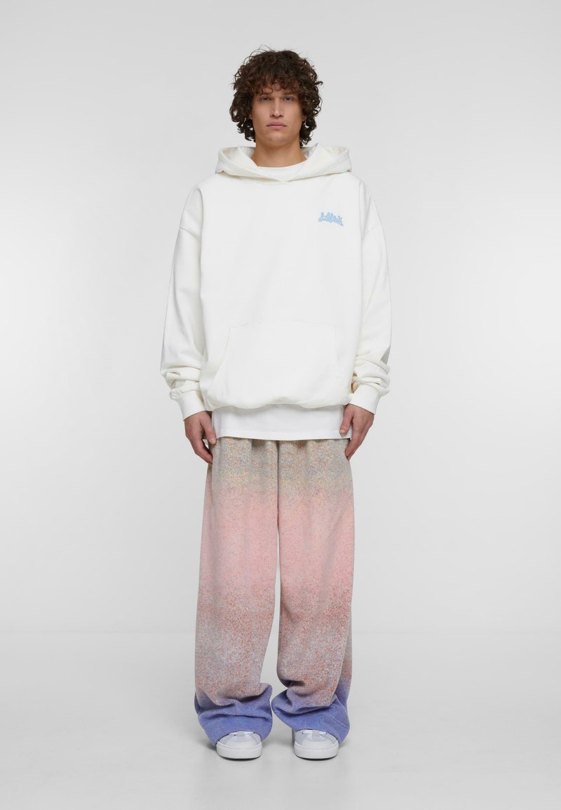 Felpa bianca con tasca frontale e logo azzurro; pantaloni larghi con un gradiente che va dal rosa al viola chiaro; sneakers bianche.
