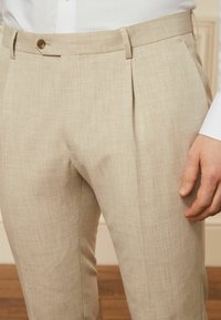 Beige maßgeschneiderte Hose aus strukturiertem Stoff, mit Falten und einem Knopfverschluss am Bund. Dezenter Glanz und gerader Schnitt.