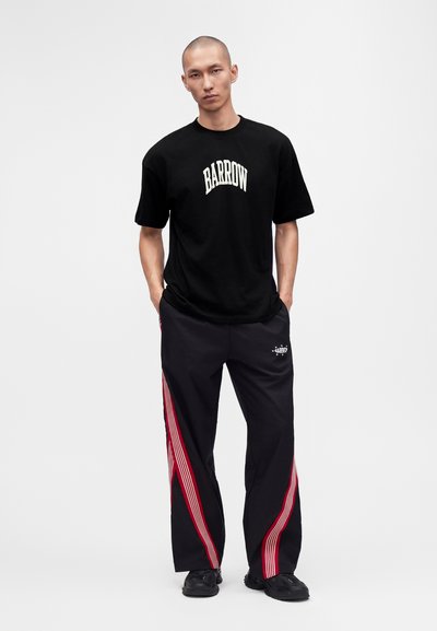 Άνδρας που φοράει μαύρο T-shirt Barrow και μαύρο παντελόνι με κόκκινες ρίγες, στέκεται με τα χέρια στις τσέπες μπροστά σε απλό φόντο.