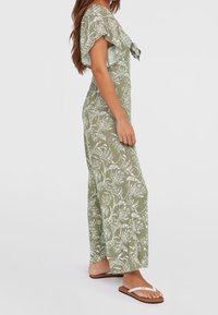 Jumpsuit verde con estampado floral, de mangas cortas, con lazo en el frente y piernas anchas. Tela texturizada, combinada con sandalias blancas simples.