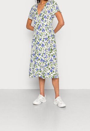 Kvinde iført en hvid midi slå-om-kjole med blå og grøn blomstret print, korte ærmer, stylet med hvide chunky sneakers, stående foran en hvid væg.