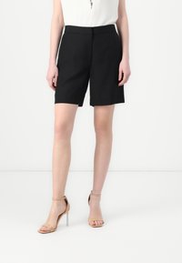 Sorte skreddersydde shorts laget av lett materiale, som går til kneet med en jevn tekstur. Parret med nude hæl sandaler med klare hæler.