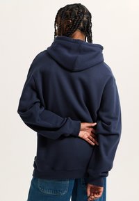 Felpa blu navy con una vestibilità rilassata, dotata di un ampio cappuccio e polsini a coste. Texture morbida e dettaglio del logo discreto sull'orlo.
