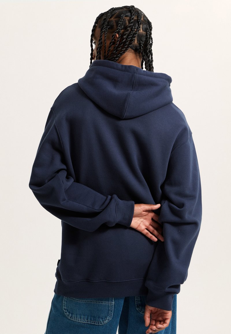Granatowy hoodie o luźnym kroju, z dużym kapturem i ściągaczami na rękawach. Miękka faktura i subtelny detal logo na dole.