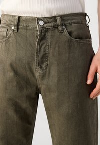 Olive green denim jeans with vertical stitching details. Features a metal button closure, five pockets, and a straight-leg cut.