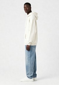 Felpa con cappuccio color crema, dotata di tasca a marsupio, abbinata a jeans azzurri chiari e sneakers bianche, esposta di lato su uno sfondo neutro.
