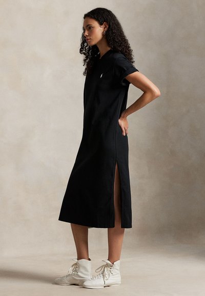 Μαύρο φόρεμα μέχρι το γόνατο από απαλό ύφασμα με κοντά μανίκια που έχουν γυριστά άκρα και πλευρικές σχισμές. Φορεμένο με λευκά μποτάκια με κορδόνια.