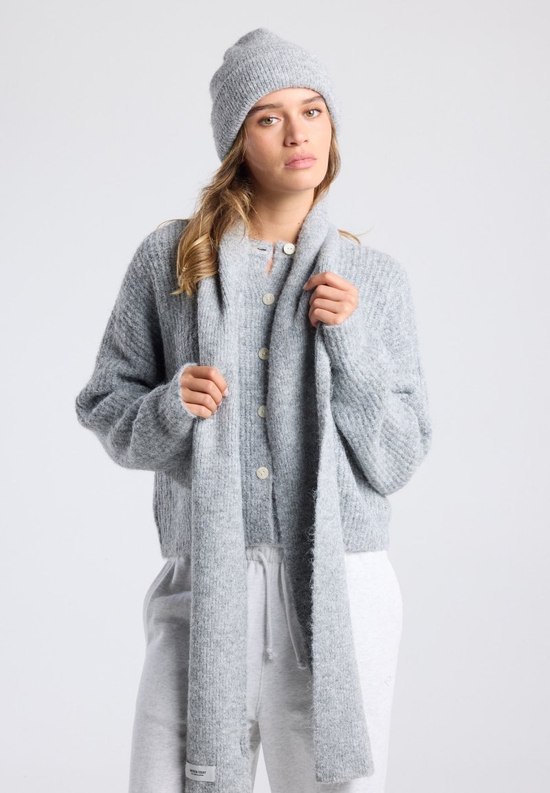 Lichtgrijze gebreide cardigan met geribbelde textuur, knoopsluiting en bijpassende sjaal. Model draagt een grijze beanie en is gestyled met casual grijze broeken.