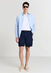 Camicia button-up azzurro chiaro sopra una t-shirt bianca, abbinata a pantaloni cargo blu navy e scarpe slip-on beige, con occhiali da sole neri.